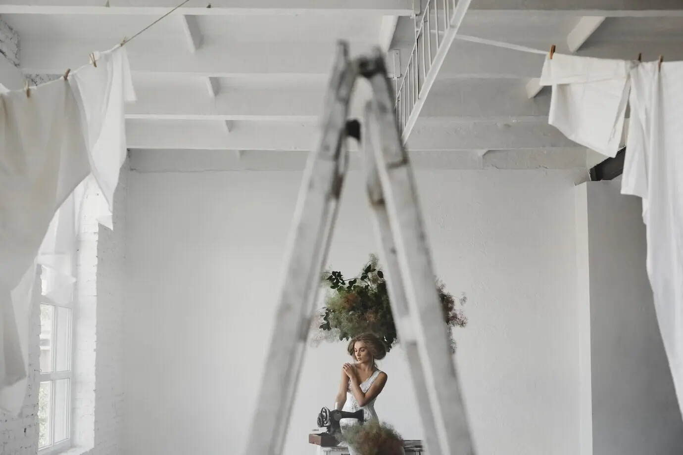 A woman wearing white sits in a room with flowers and a sewing machine.