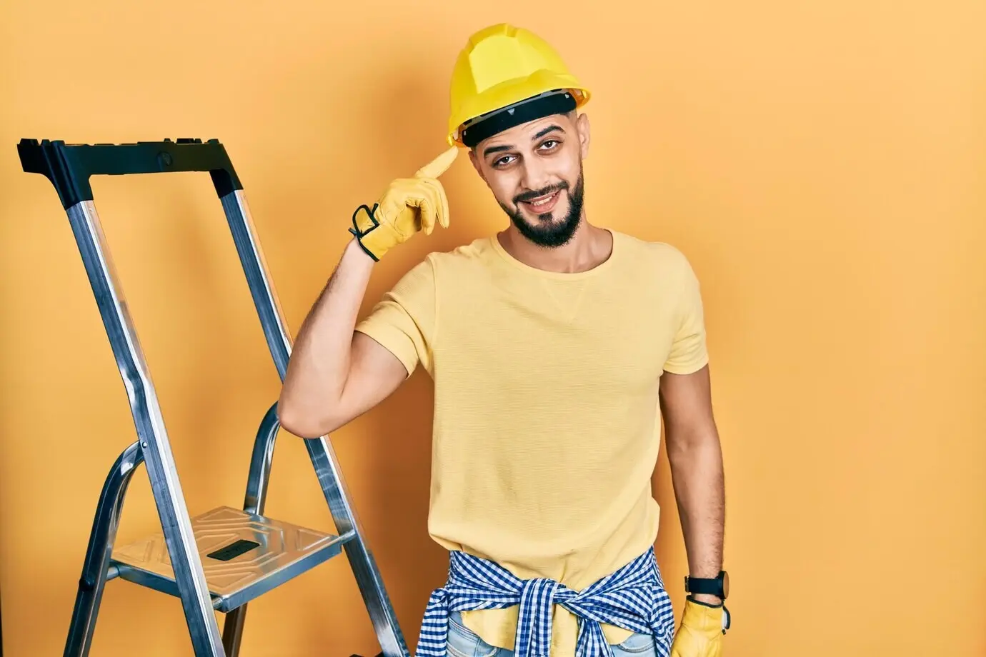 A handsome bearded man by construction stairs, wearing a hard hat, smiling and pointing at his head with one finger, indicating a great idea or thought or a good memory.