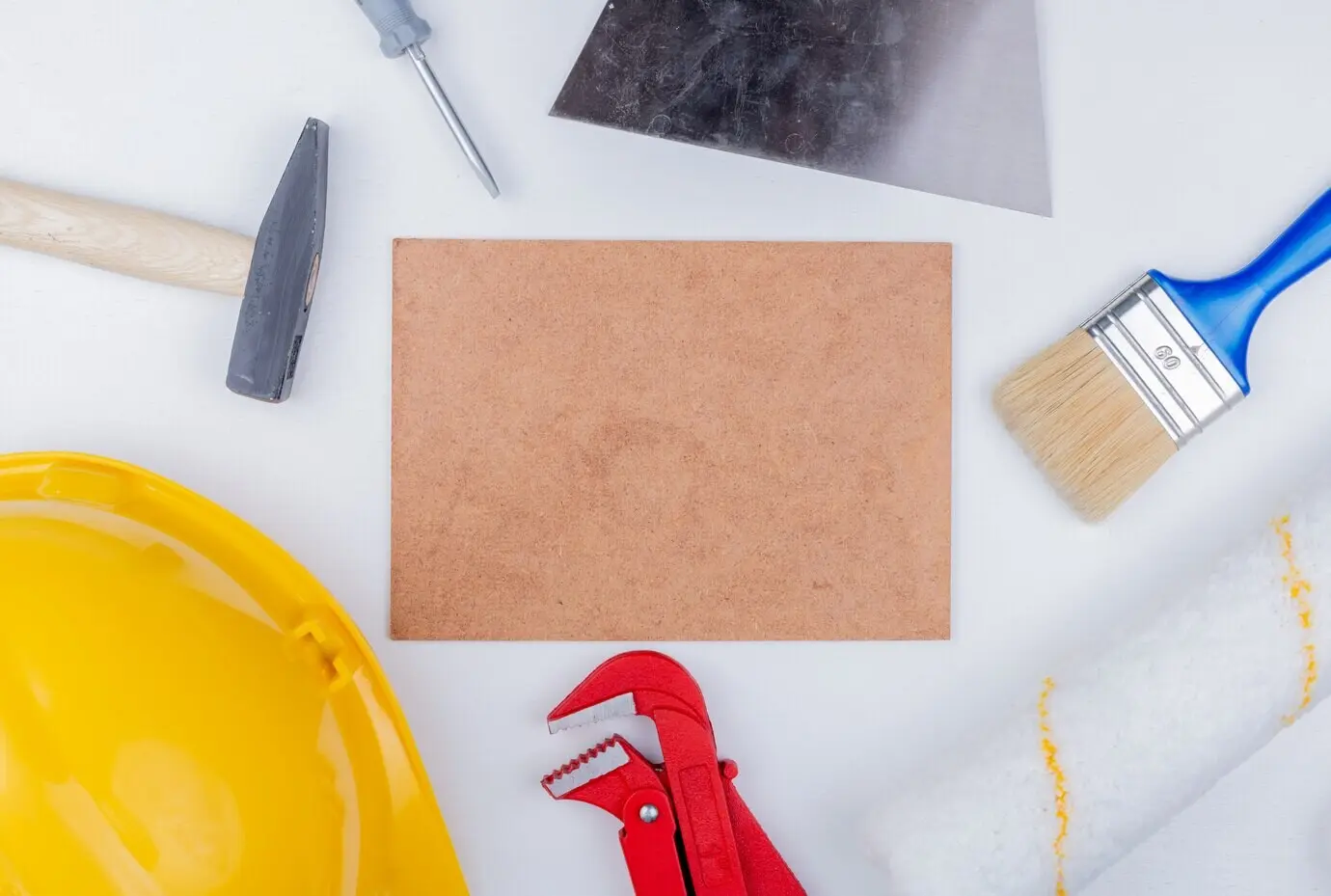 Top view of construction tools, including a brick hammer, safety helmet, screwdriver, pipe wrench, paint brush and roller, and a putty knife, arranged around a mettlach tile on a white background.
